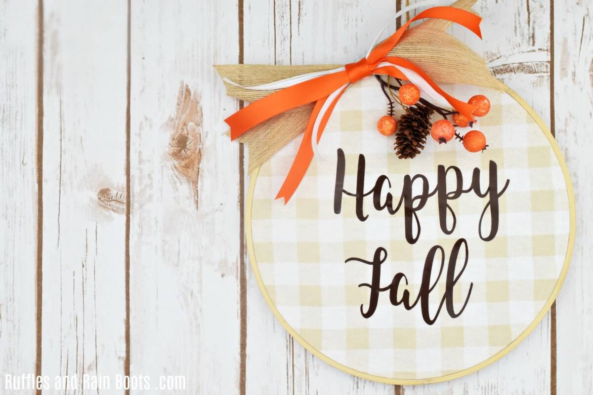 Horizontal image of a large fall wreath idea of Buffalo check fall wreath with free happy fall SVG cut file, farmhouse bow, and fall embellishments hanging on a white wood plank wall. 