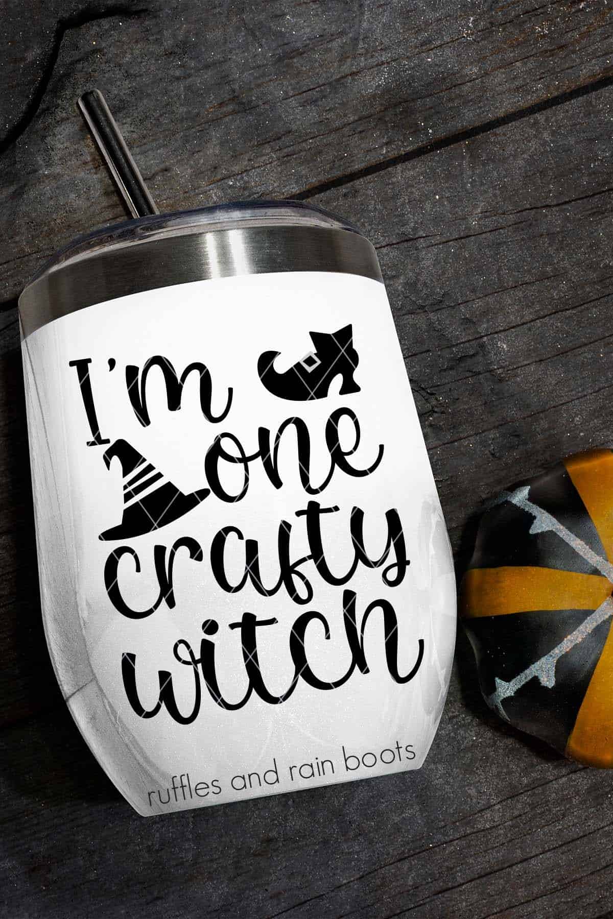 Vertical image of a white tumbler with I'm One Crafty Witch SVG done in black vinyl with a witch hat SVG and a shoe cut file on black wood.