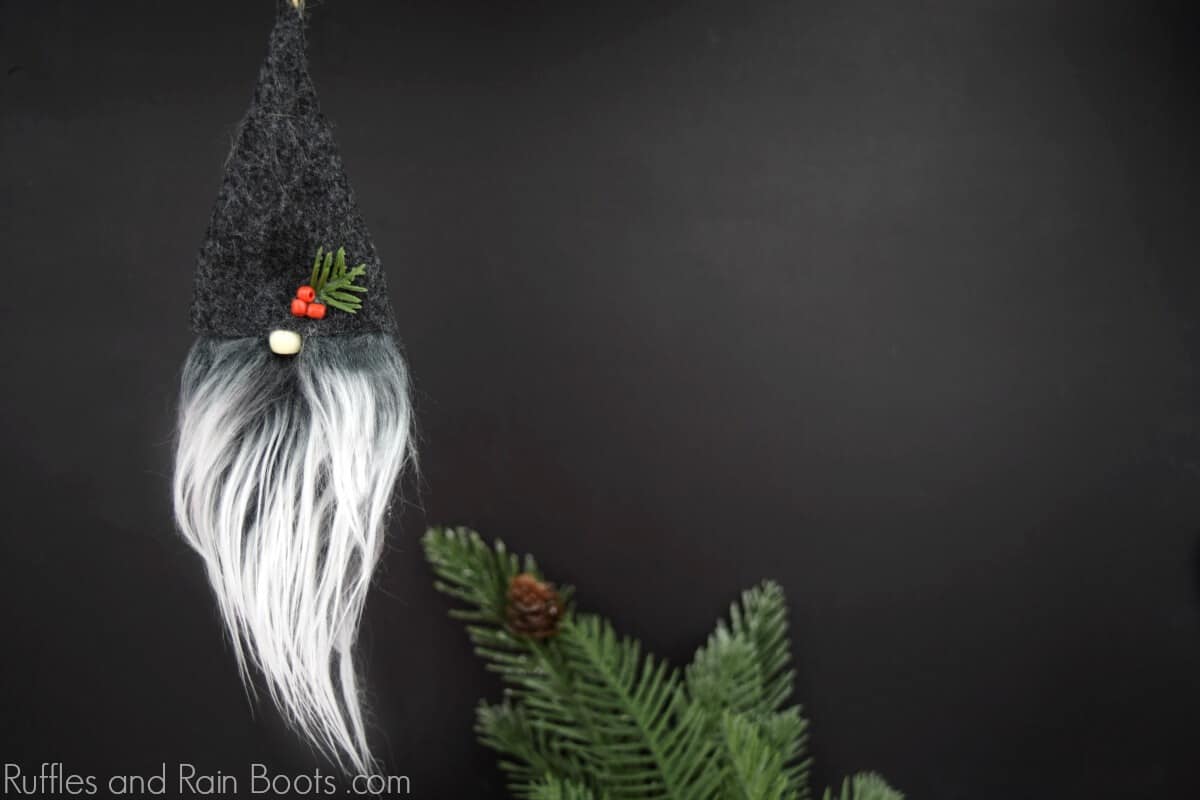 Horizontal image of a gray hat, white and gray beard, and beaded gnome ornament hanging over pine branches on black background.
