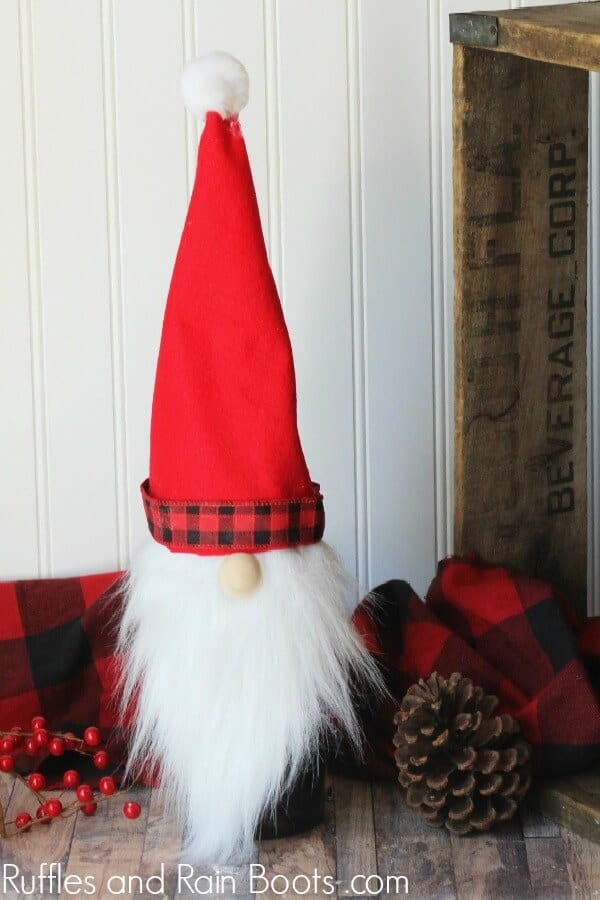 A tall Scandinavian gnome wine bottle topper with a red hat and white beard on wood background with festive holiday towel, berries, and pine cone. 