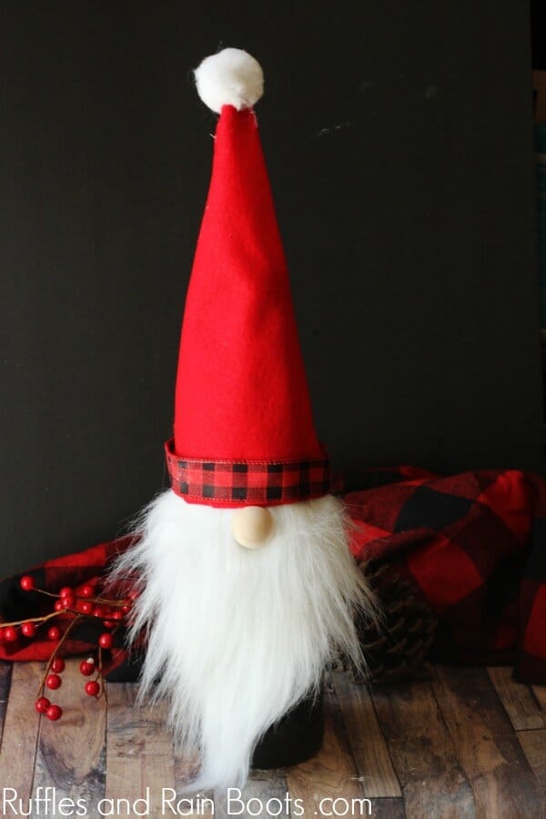 Vertical image of a Christmas gnome wine bottle topper with a red hat, brim, and white beard on a dark background.