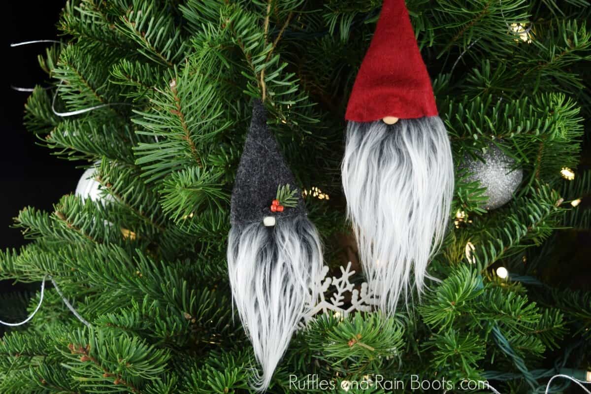 Horizontal image of two no sew gnome ornaments hanging on a lighted Christmas tree with felt hats and Mongolian fur gnome beards.