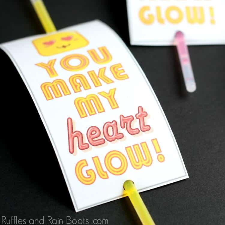 Square image of a glow stick Valentine idea on a black background with a yellow glow stick and a white card which reads you make my heart glow.