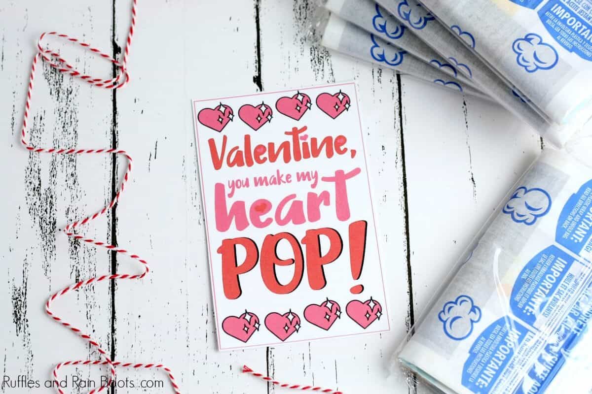 horizontal image showing red and white twine, bags of microwave popcorn, and a printable card which reads Valentine, you make my heart pop all on a white wood background