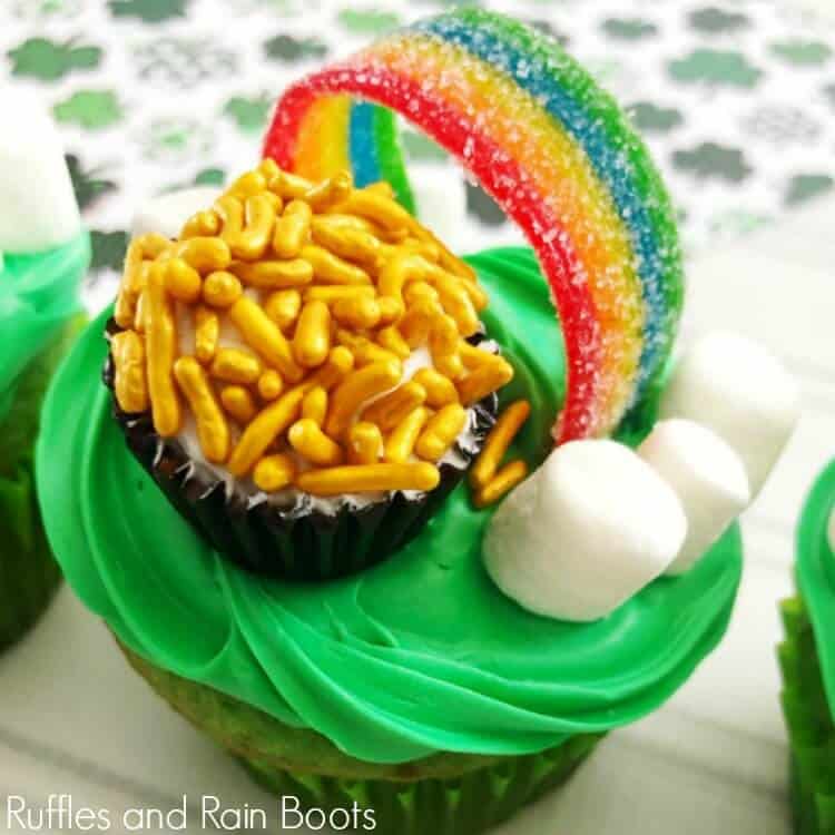 square close up image of a rainbow candy, mini marshmallow clouds, and a Ferraro Rocher turned into a pot of gold all on top of a cupcake with green frosting