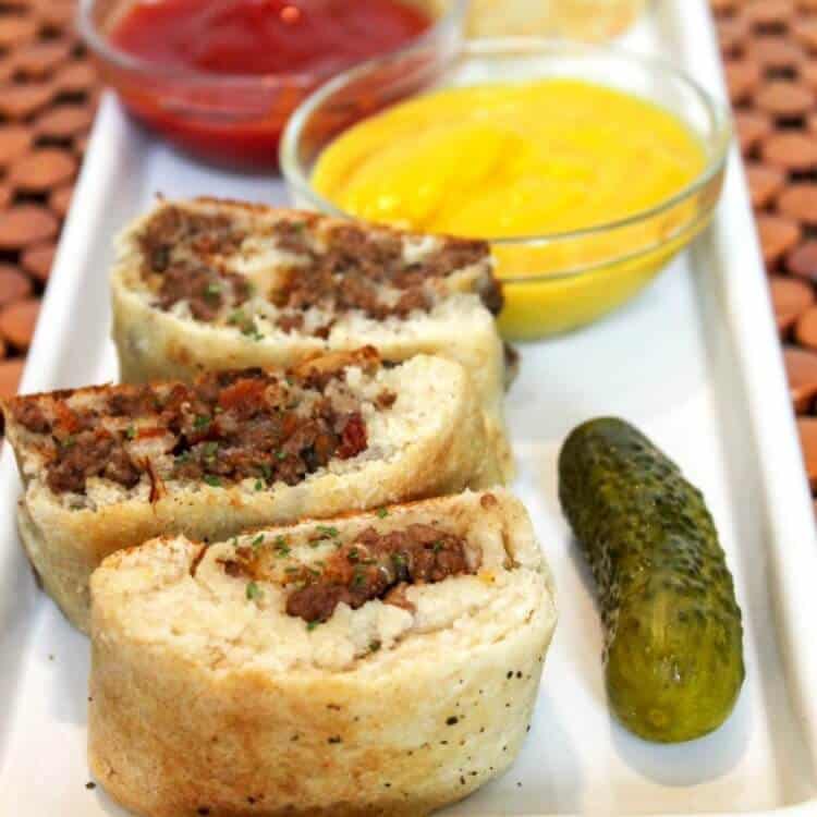 side view of cheeseburger pinwheels on a white plate with a pickle, a bowl of mustard and a bowl of ketchup on a red and black checked background