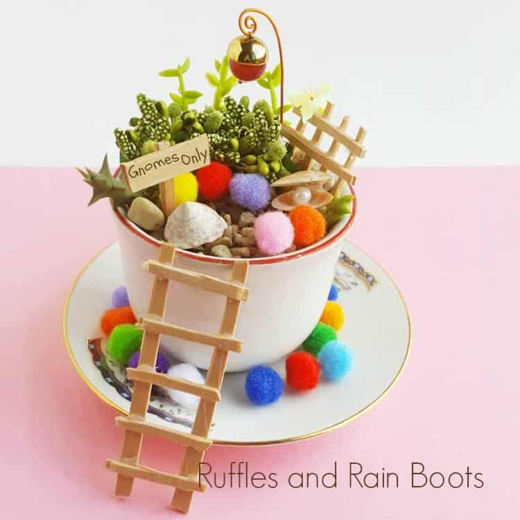 Square image of a teacup full of pompoms, plants, wooden signs, and shells sitting on a pink table.