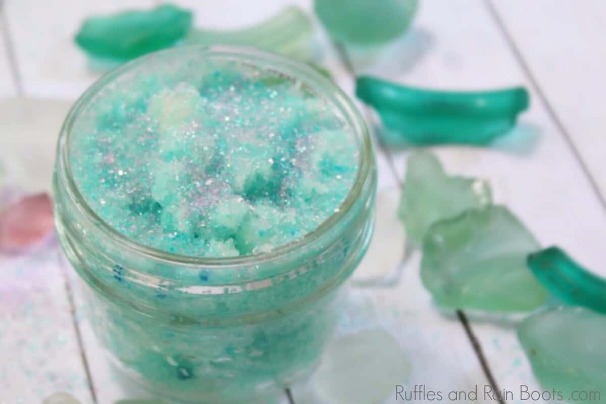 teal mermaid sugar scrub in a clear jar on a white background surrounded by teal and green sea glass pieces