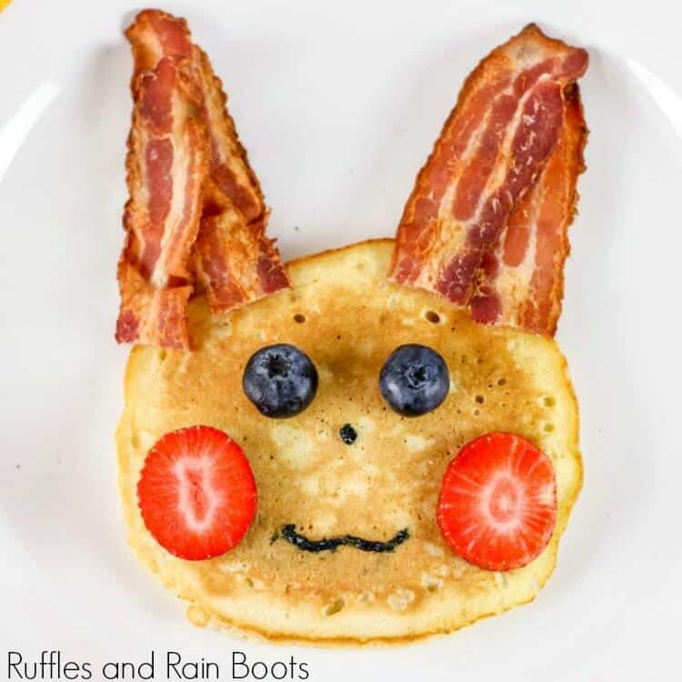 pancake with bacon "ears", strawberry "cheeks", blueberry "eyes", and icing for a nose and mouth made to depict a pikachu from pokemon on a white plate 