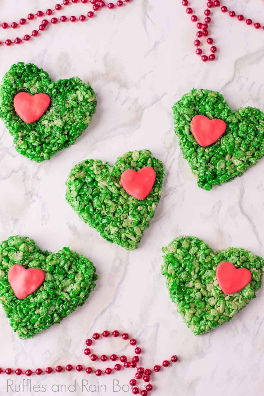 green grinch rice krispies treats with candy hearts on white marble background with red beads and no text.