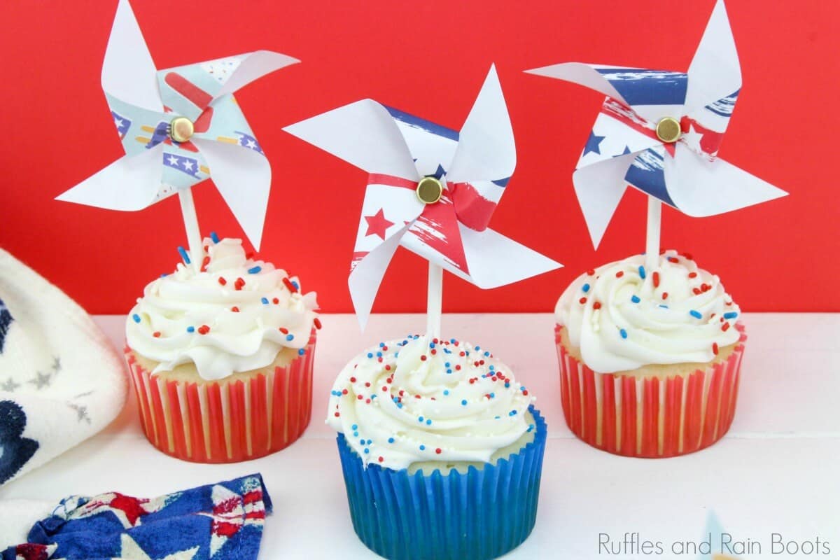three simple 4th of July cupcakes with white icing on top and red and white cupcake liners with pinwheel toys on top on a white table with a flag linen and a red background
