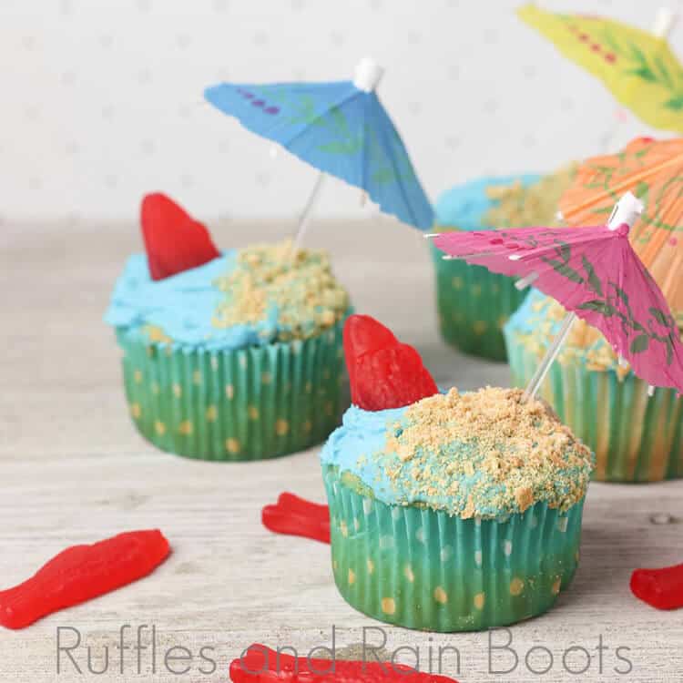 blue frosted cupcakes with green cupcake papers with tiny umbrellas and red candy fish sticking out of blue frosting on a light beige background with scattered red candy fish
