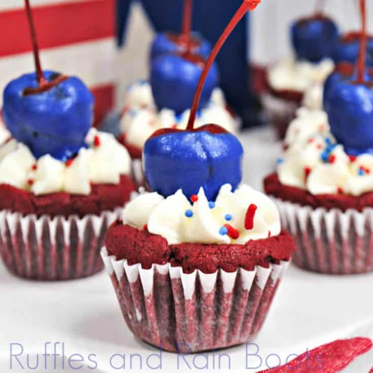 side view of several red velvet brownie bites, red brownie mini-cupcakes with white icing topped with blue-chocolate covered cherry on top on a white plate with a flag background