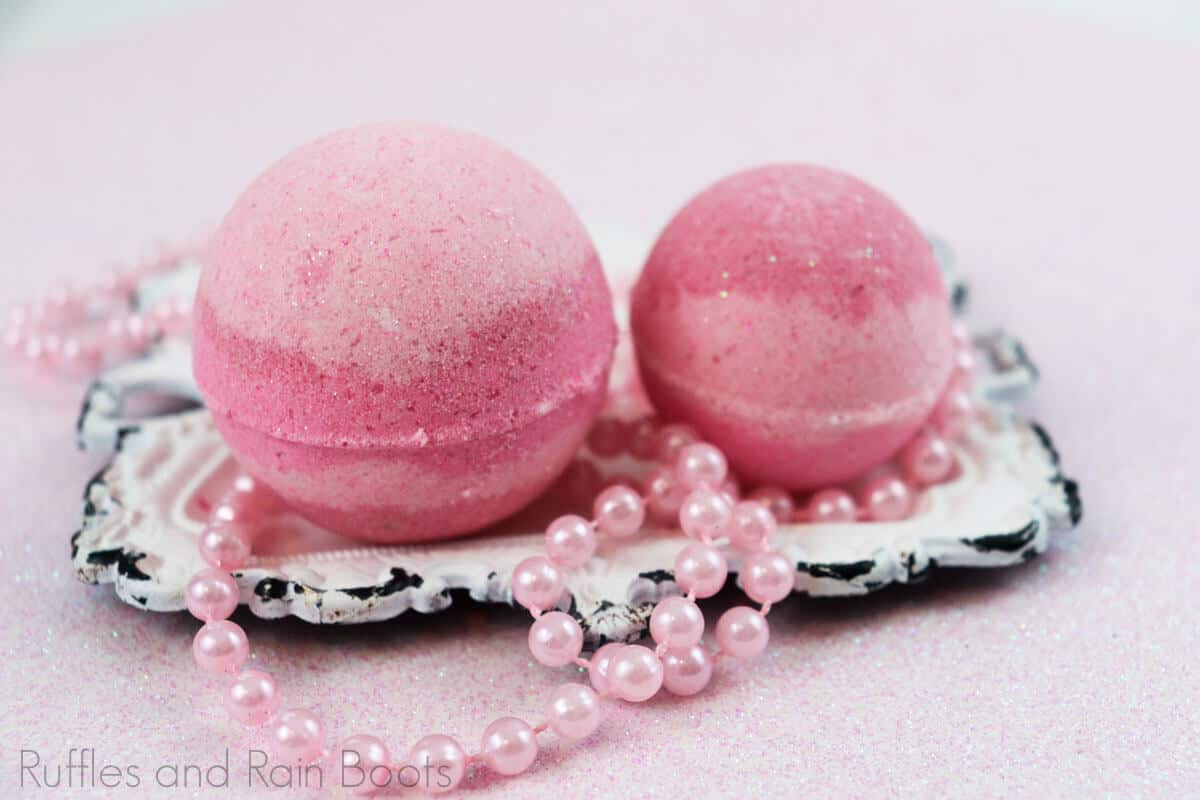 Close view of two pink striped princess aurora bath bombs on a white antique square plate with toy pink beads on a pink table.