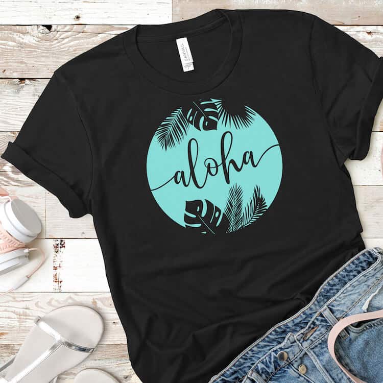 Square image of black t shirt with teal aloha free summer SVG vinyl with jean shorts and headphones on a beige wood background.