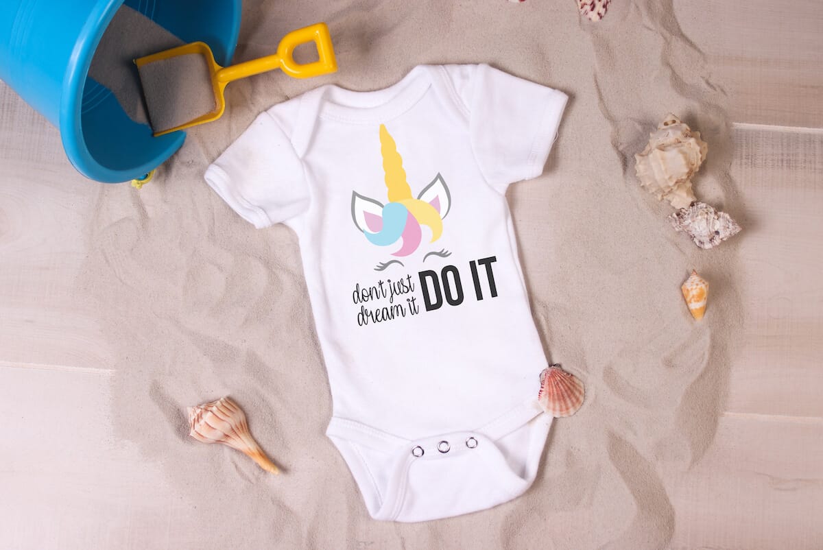 Don't Just Dream It Do It free unicorn SVG on baby onesie in a bed of sand with a toy shovel and pail and sea shells scattered around