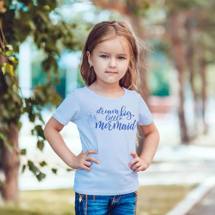 Dream big little mermaid cut file in navy glitter on a light blue t shirt worn by a young girl in jeans photographed in a park with no text.