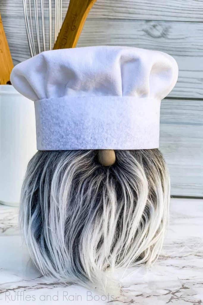 close up of chef gnome with chef toque, gray mongolian fur beard, and polymer clay nose on white wood and marble background with baking utensils
