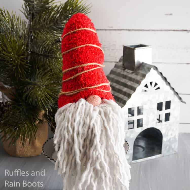 Square image of a mop beard gnome made with dollar store materials on gray wood table with mini Christmas tree and metal house.