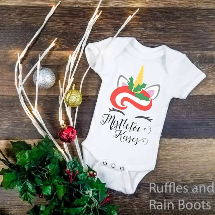 mistletoe kisses unicorn christmas svg on onesie on a dark wood background with a holiday swag