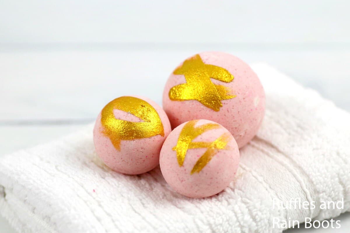Three pink and gold painted bath bombs on a white bath towel on white background.