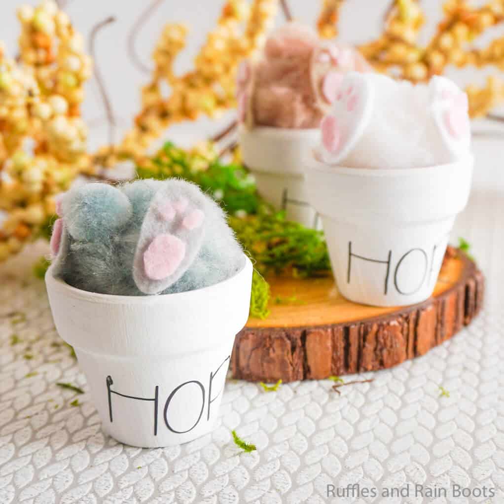 Vertical image of a quick Easter bunny butt craft in clay pot with Rae Dunn inspired writing placed on a textured table with wood round and flowers.