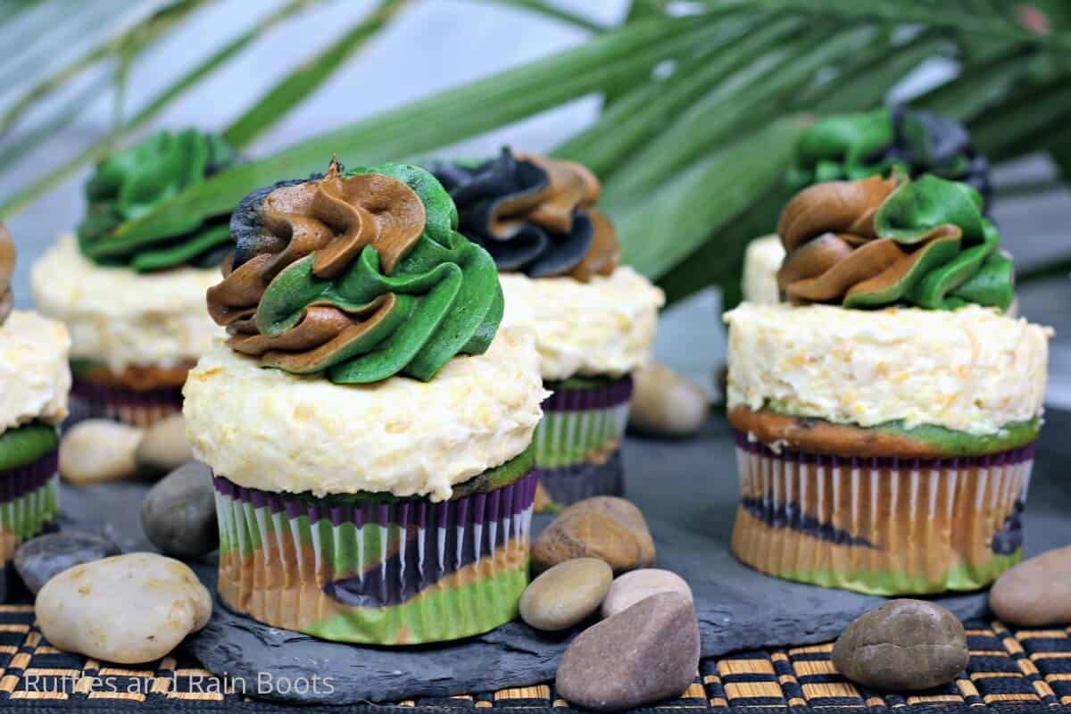Horizontal image showing camouflage cupcakes, cereal frosting, and three color frosting topper in camo on a bed of slate with rocks for a jungle or safari themed party.