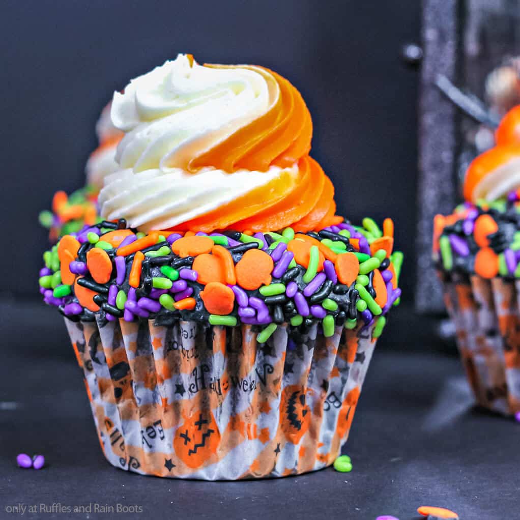 Square image of an orange and white frosted chocolate cupcake with a bed of sprinkles and Halloween cupcake wrappers in front of a black background.