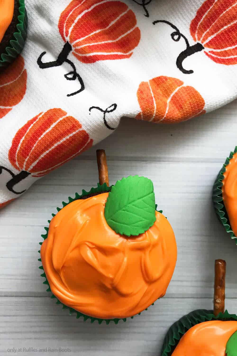 overhead view of an orange frosted chocolate cupcake with a pretzel stem and a green fondant leaf on a white wood background with a fall kitchen towel