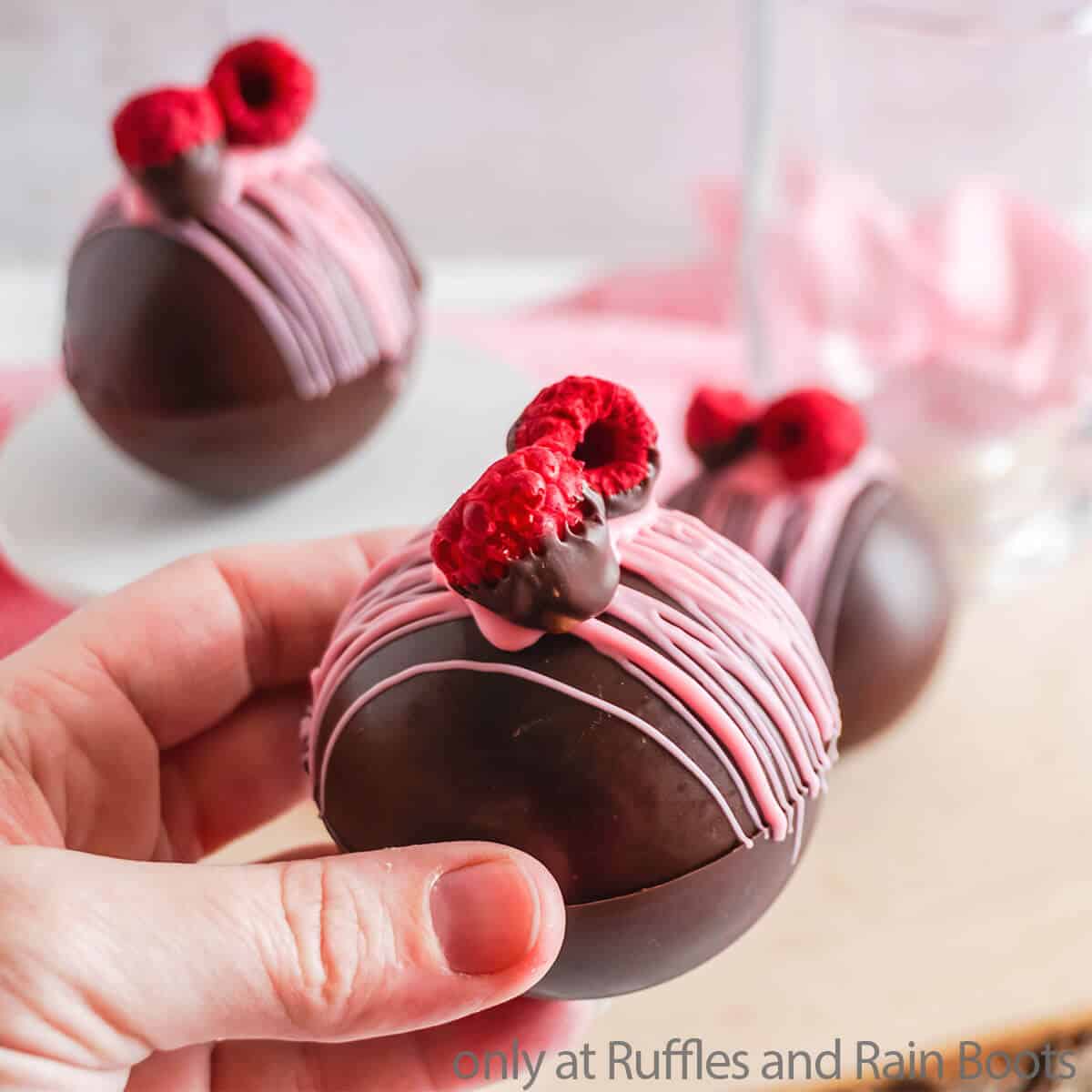 Close up of a hand holding a raspberry dark chocolate hot cocoa bombs on white and pink background.