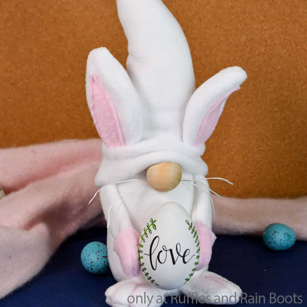 Square image of a white Easter bunny gnome holding an egg in front of a cork background.