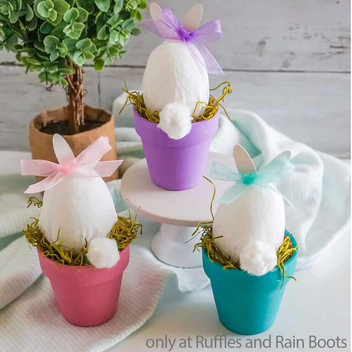 Square image of three bunny in a pot easter crafts painted pink blue and purple staged on a white table with a miniature tree in a rustic paper bag pot.