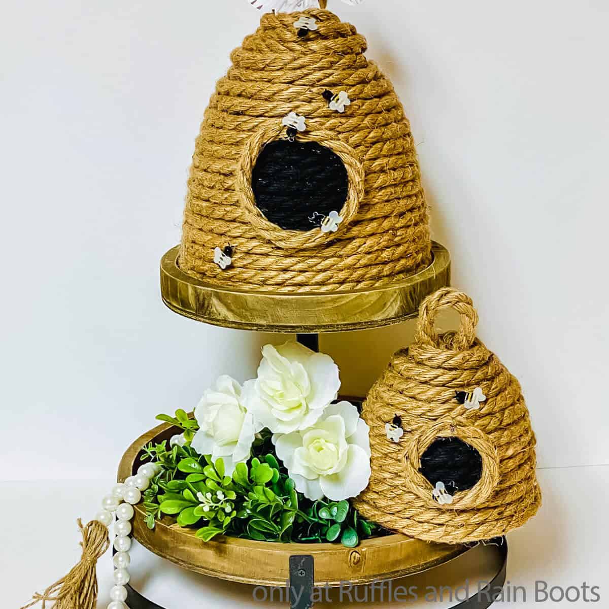 Square close up image of two sizes of DIY beehive crafts made with rope on a tiered tray with greenery and flowers.