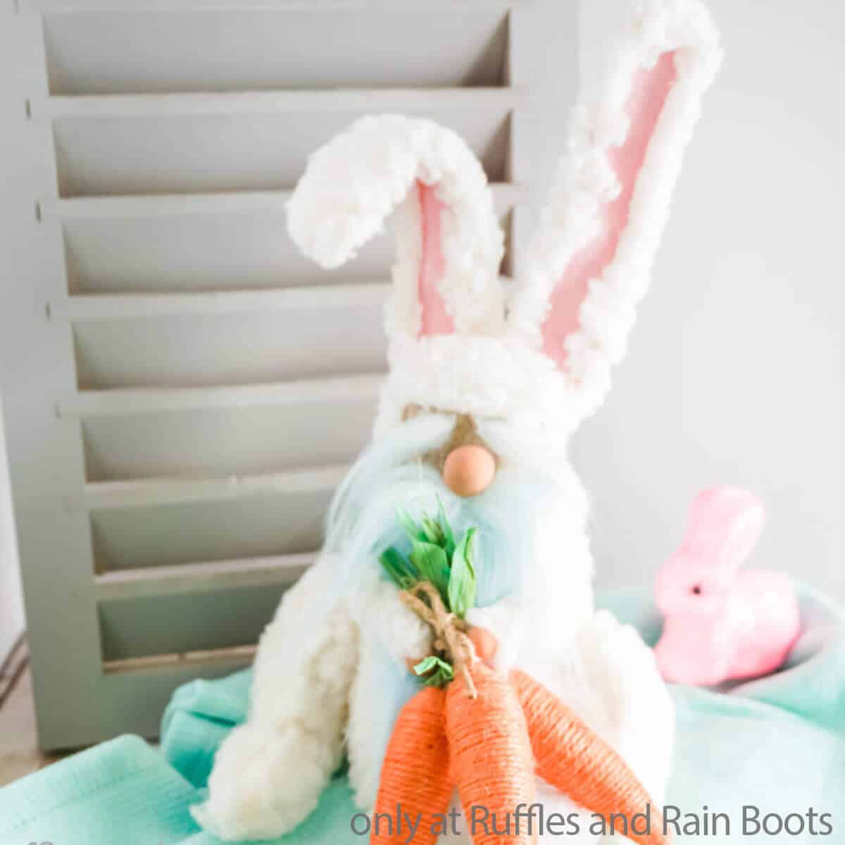 Square image of a light blue bearded Easter bunny gnome with carrots sitting in front of a shutter and an Easter themed background. 