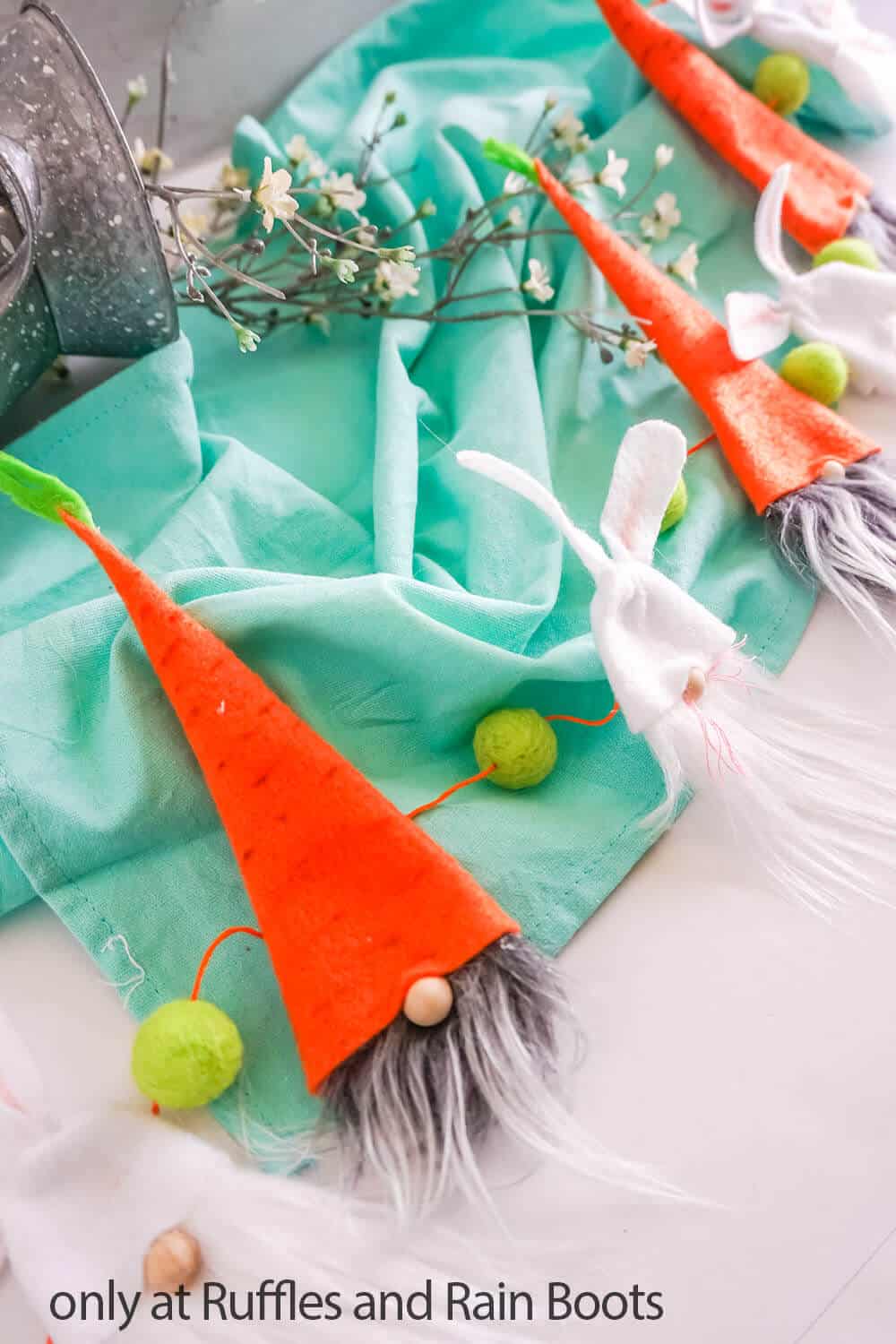 Orange carrot hat gnomes and bunny gnomes strung on a Spring garland placed on a table with teal towel and tiny flowers. 