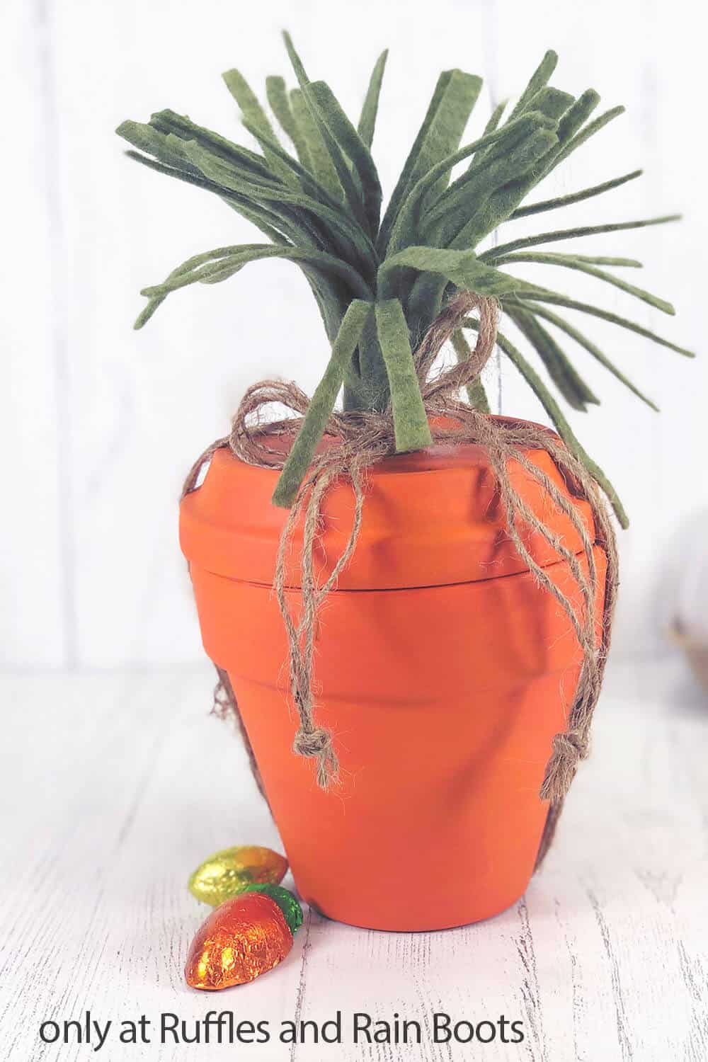 Close up image of a terra cotta pot painted orange to resemble a carrot  with twine and green felt placed on a white wood background. 