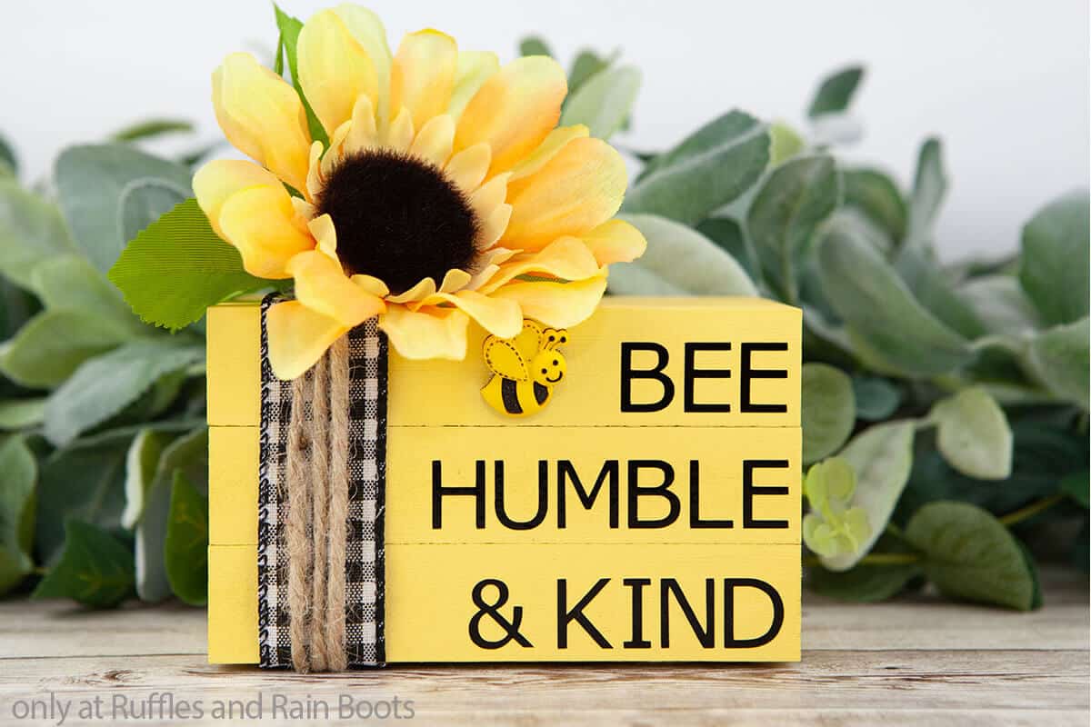 Horizontal image of a yellow bee book stack in front of lamb's ear and mixed greenery on a blonde wood talbe.