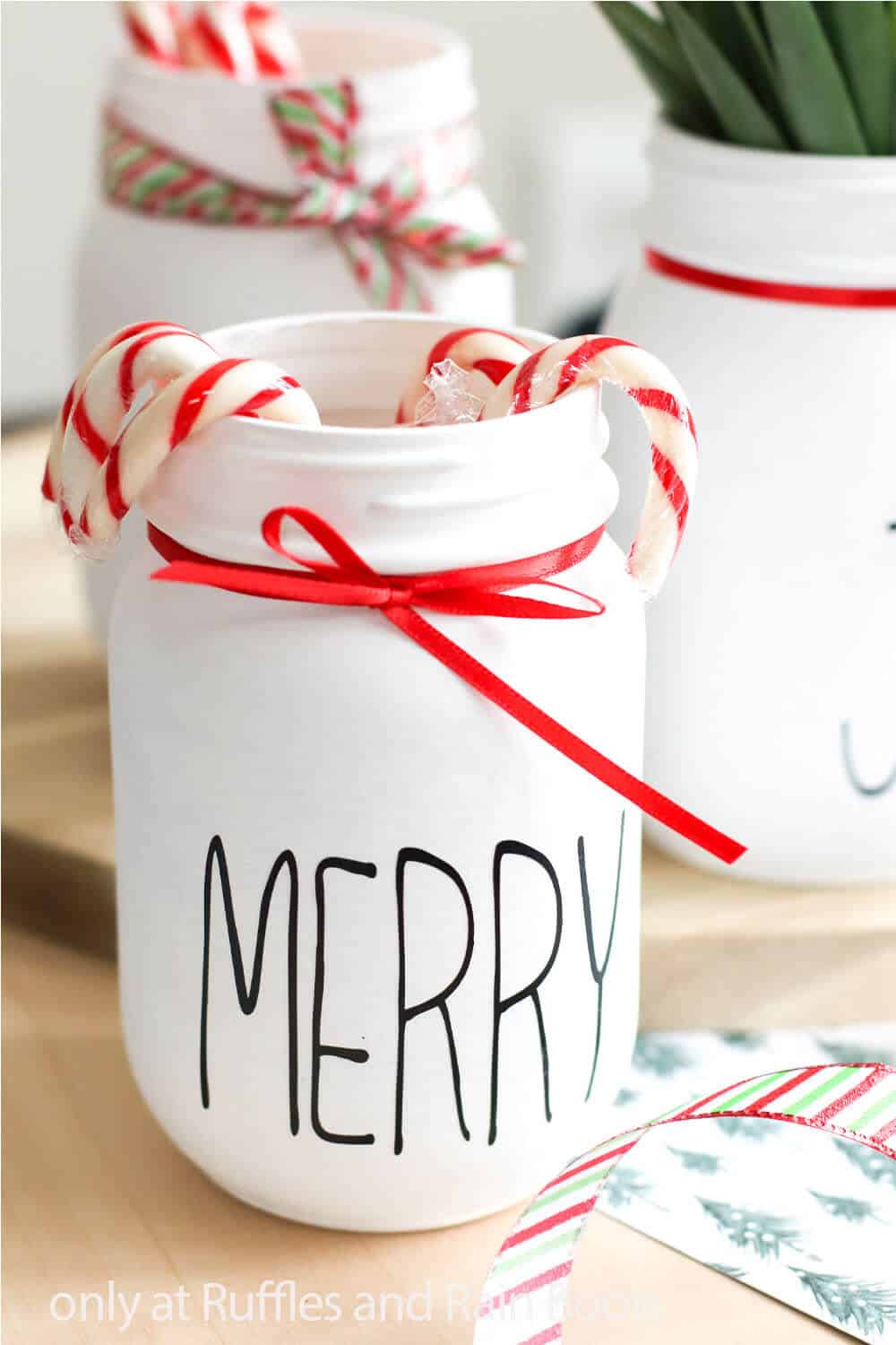 A 16 ounce white painted jar Christmas Cricut craft with vinyl text which reads merry tied with a thin, red ribbon.
