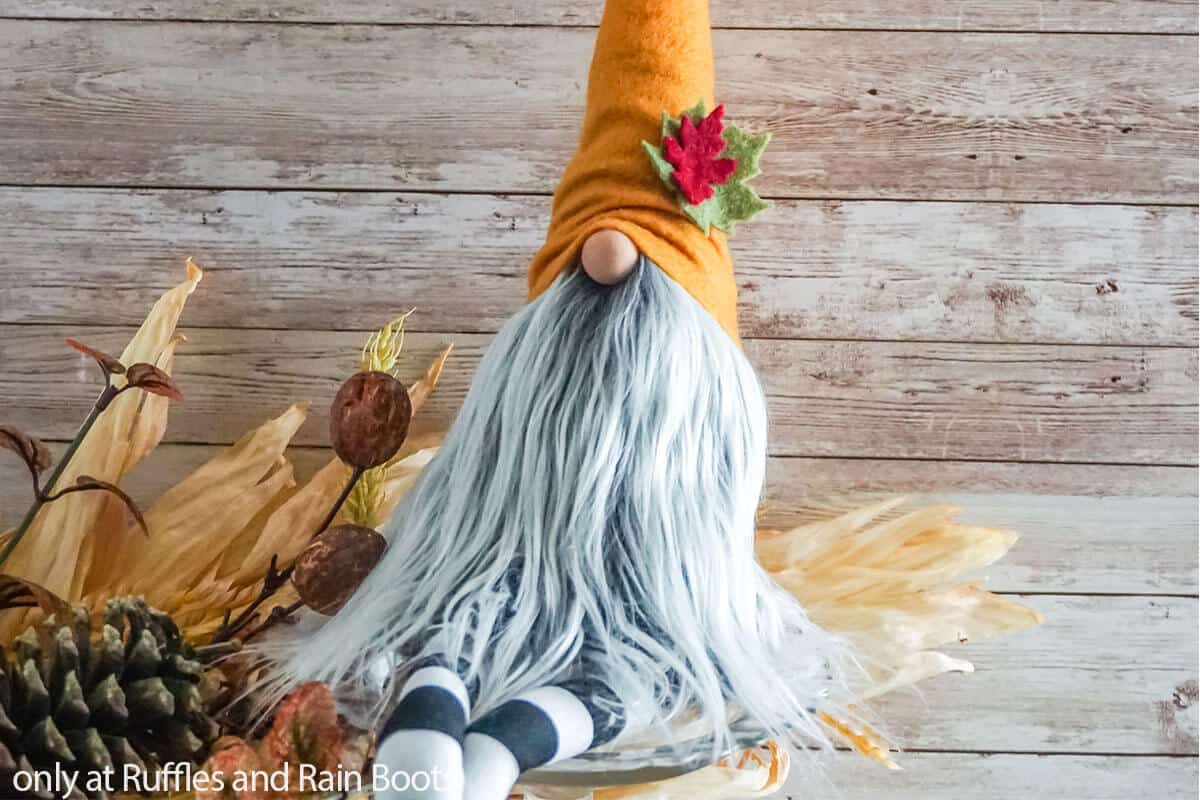 Horizontal image of a long legged gnome with dangling legs made with a free gnome pattern sitting in front of fall foliage and a wood background.