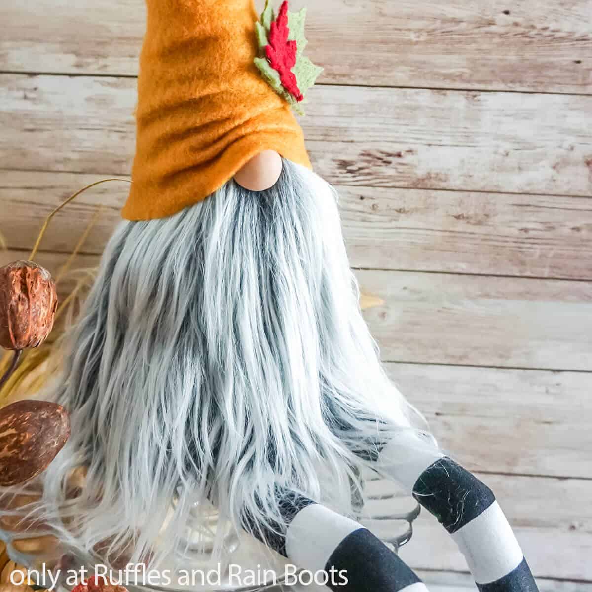 Close up image of a fall shelf-sitter gnome with gray and white beard, striped legs, and a burnt orange hat for fall in front of a wood background.