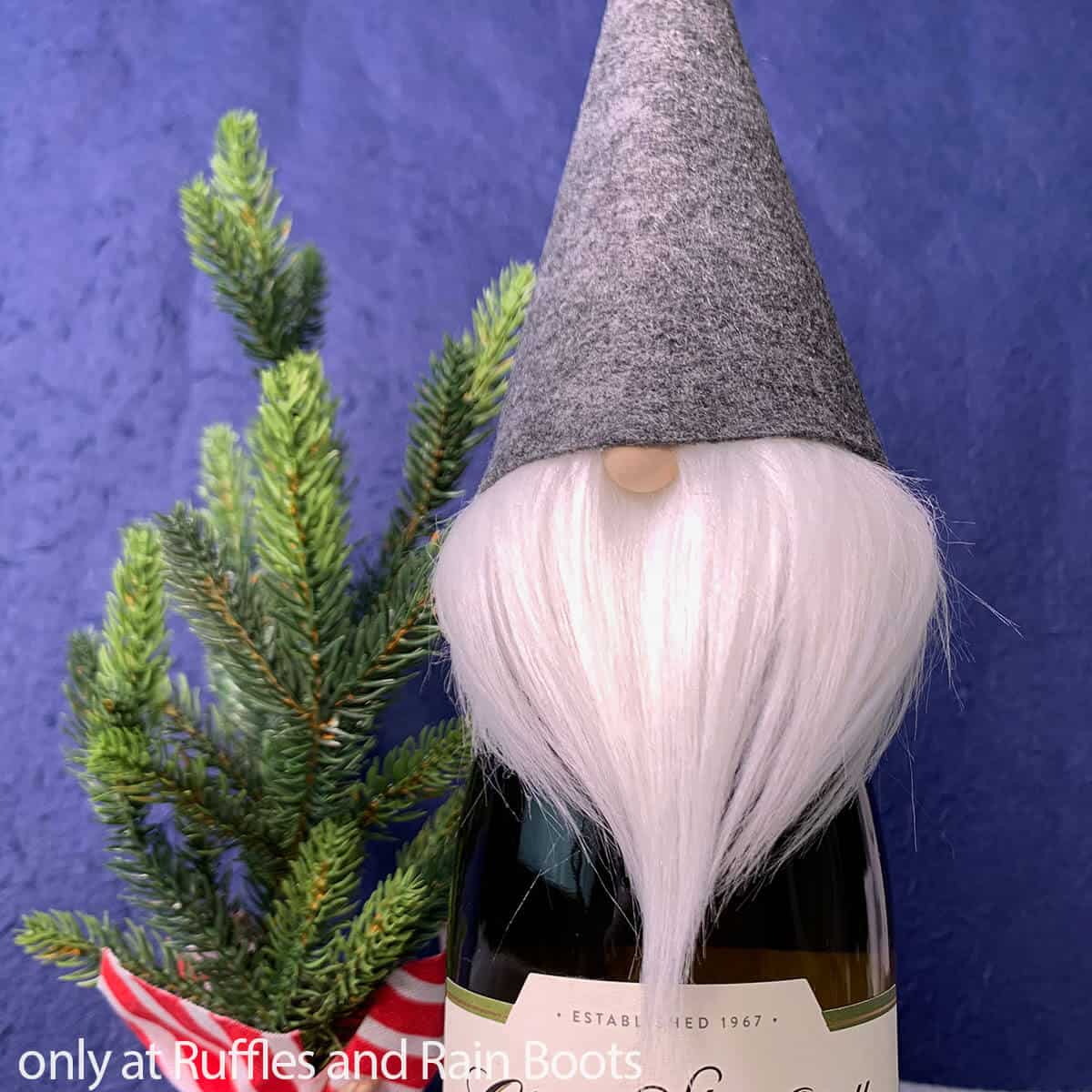 Square close up image of a gray hat felt gnome wine bottle topper with white beard on a bottle of wine next to a mini pine tree in front of a blue background.