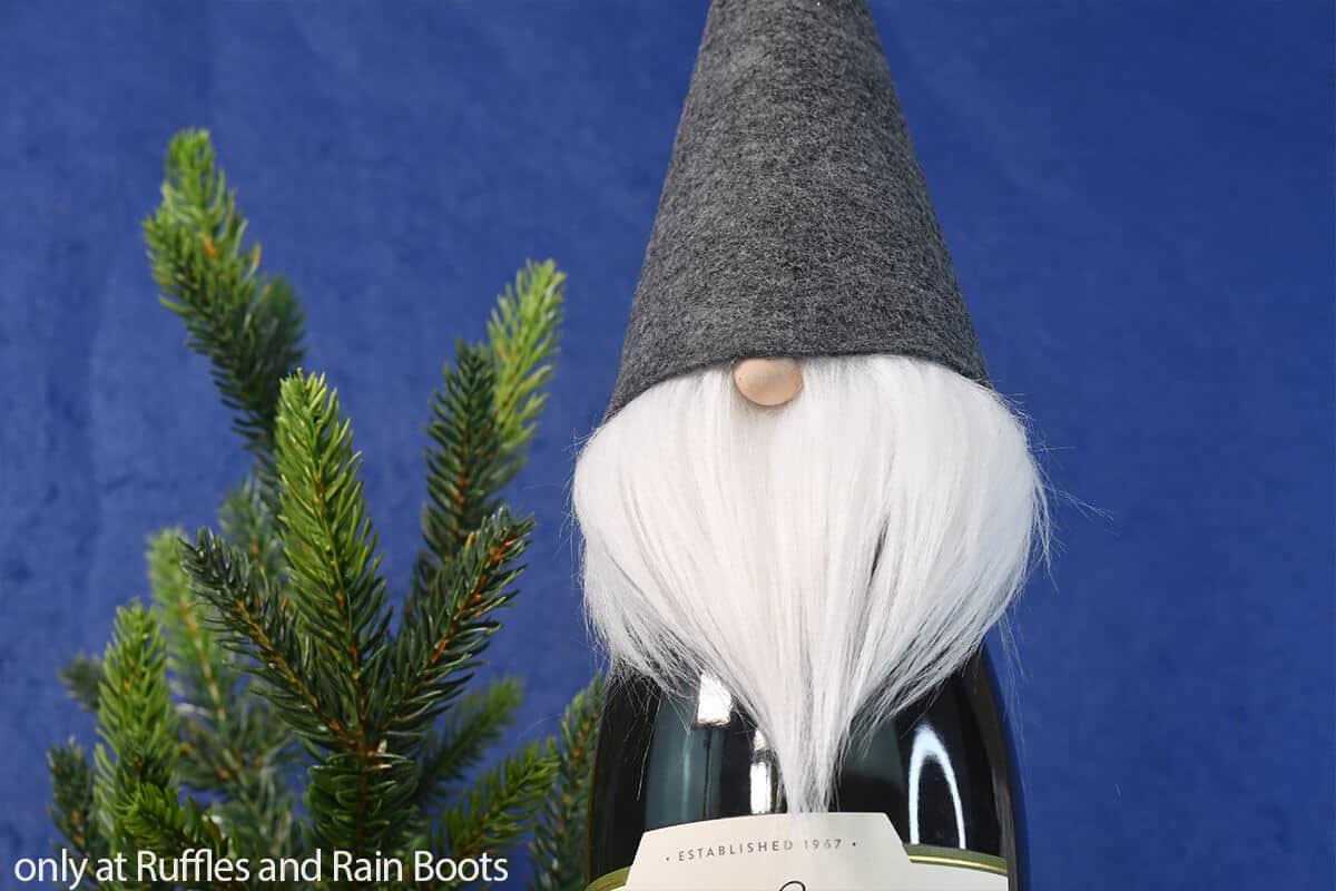 A gray felt gnome wine bottle cover on a bottle of wine in front of a small pine tree and a blue background.