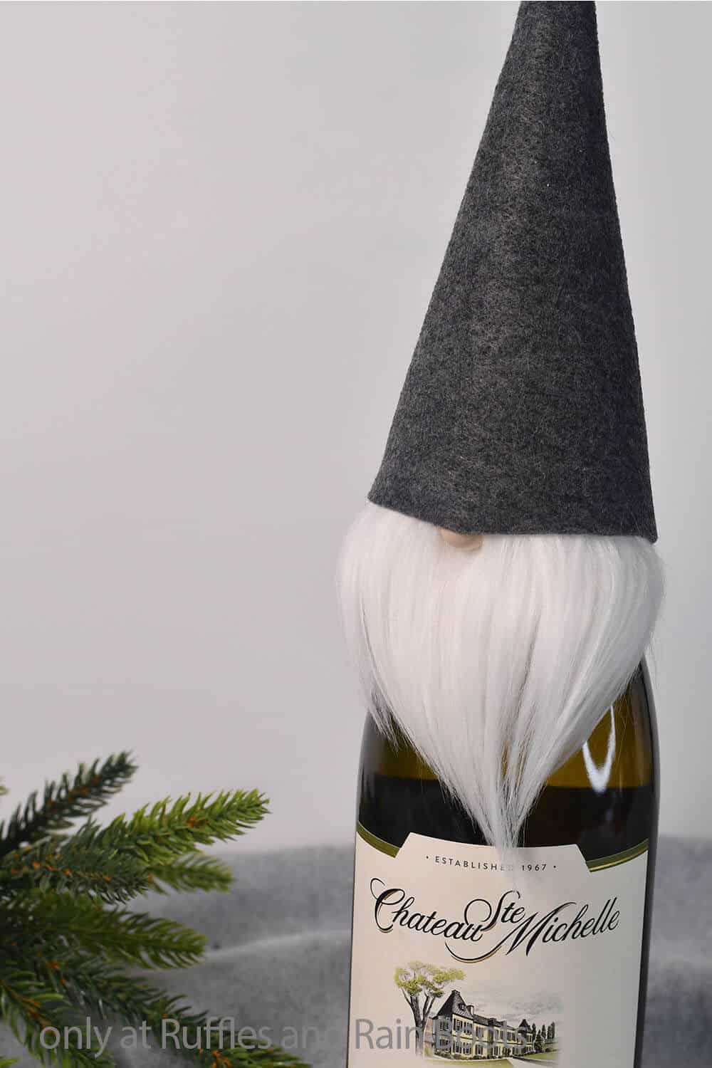 A vertical image showing a felt gnome wine bottle topper on a wine bottle next to a pine spring on a gray fleece desk and a white background.