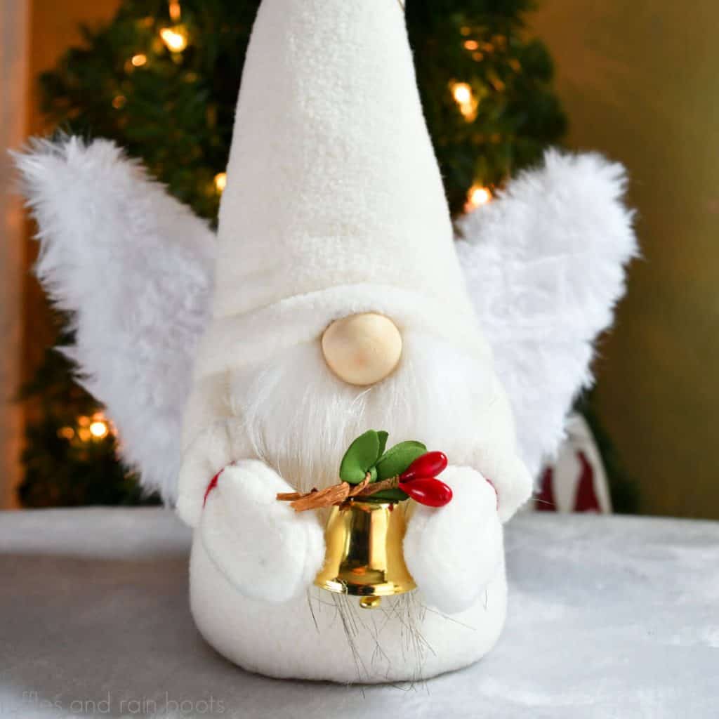cream and white angel gnome holding gold bell with Christmas greenery in front of a Christmas tree
