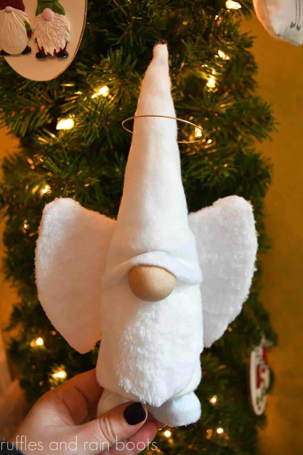 vertical image of diy angel gnome held in front of a Christmas tree with gnome ornaments and soft light