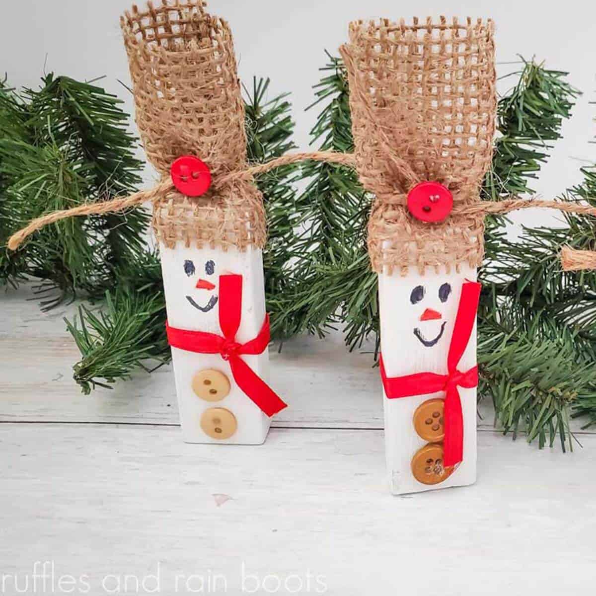 square image of 2 snowmen ornaments made with tumbling tower blocks from dollar tree painted white and given rustic burlap hat red ribbon scarf and buttons in front of Christmas greenery