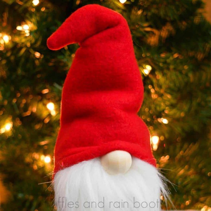 square image close up of red hat gnome with white beard and wood nose in front of a lit Christmas tree