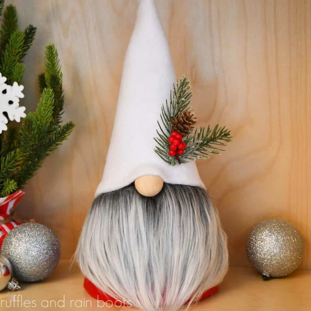 square image close up of a Christmas gnome in a white had with red body and frosted gray gnome beard with pine cone and berry accent in front of a holiday background with mini tree and silver ornaments