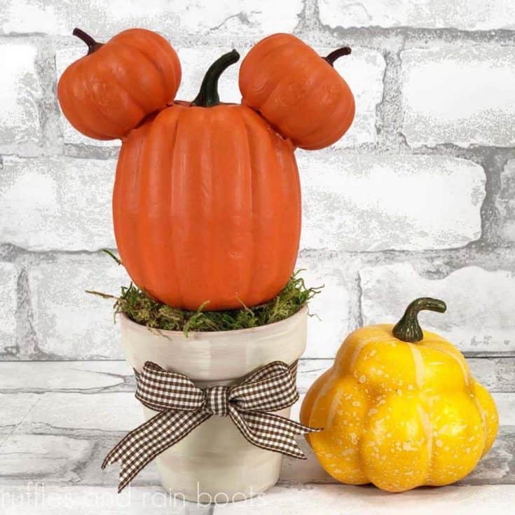 square image of pumpkin Mickey Mouse topiary on a white brick background with a yellow squash for fall