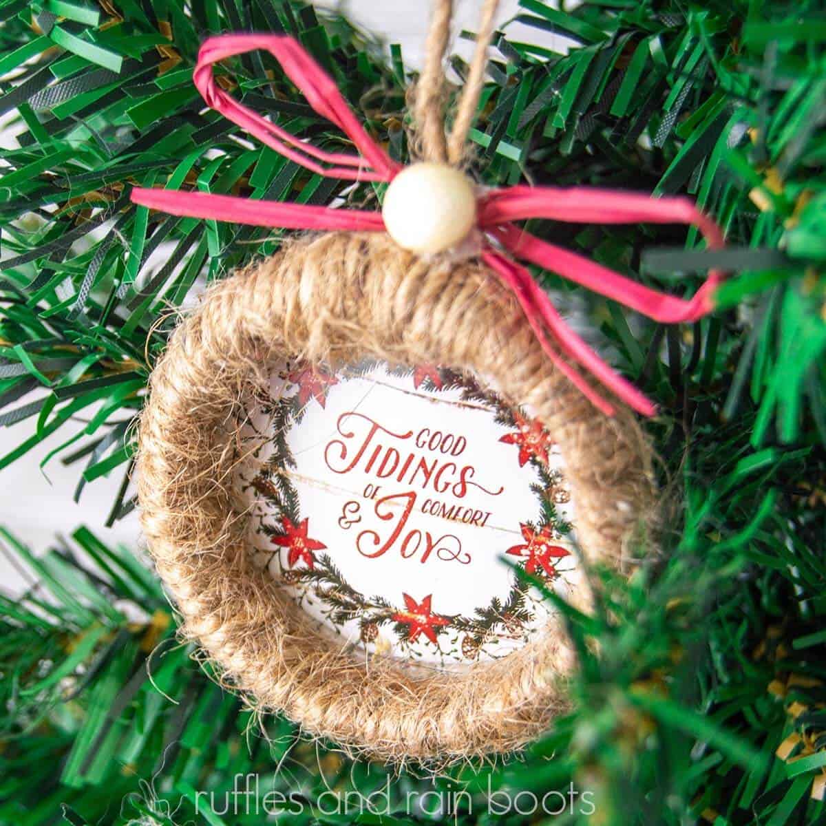 square close up image of a Dollar Tree twine wreath ornament with red bow and good tidings of comfort and joy calendar page hanging on a faux Christmas tree